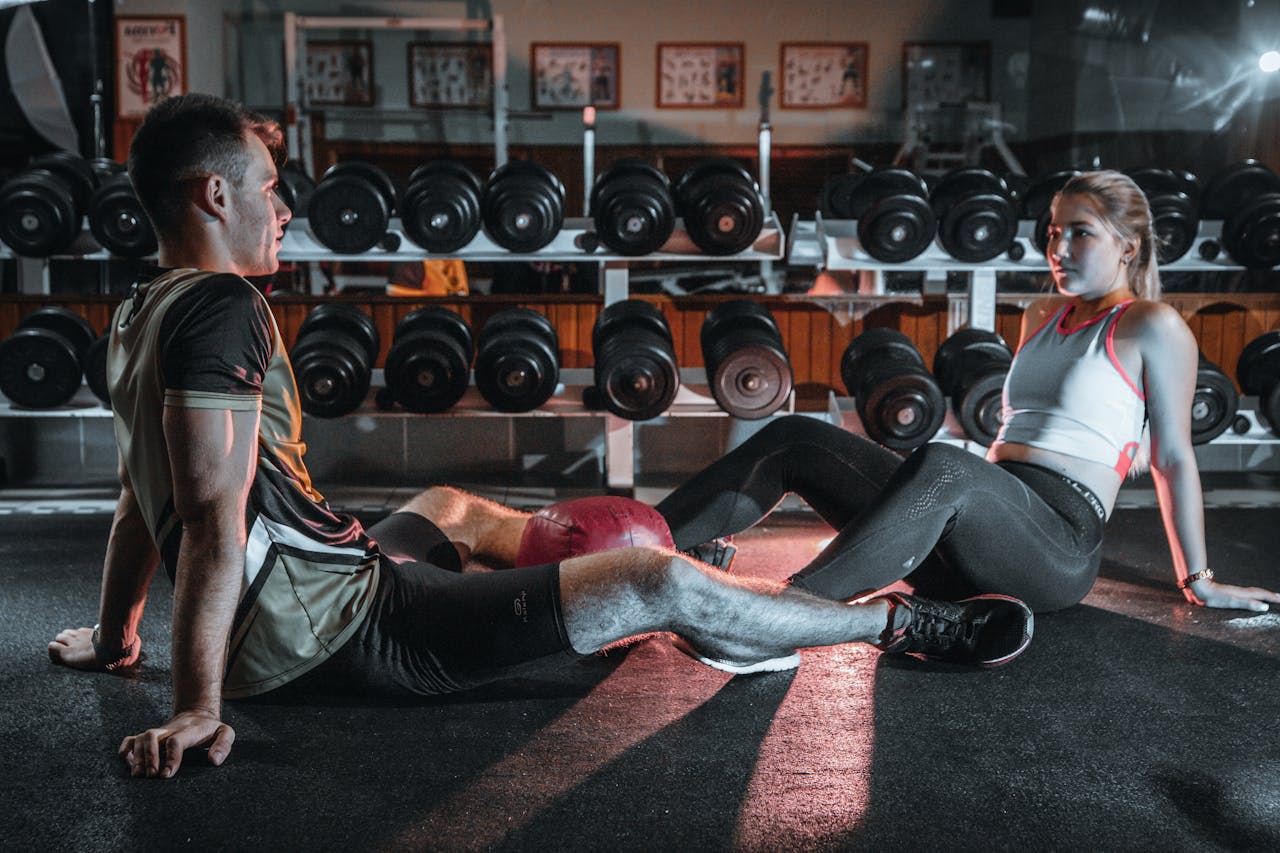 A man and woman resting together on the gym floor, surrounded by dumbbells.